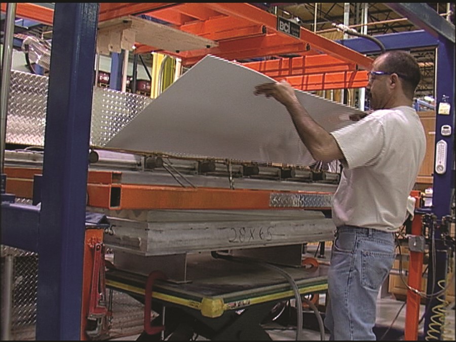 Factory worker in a white shirt and safety glasses lifts a large aluminum sheet over machinery, no visible expression.