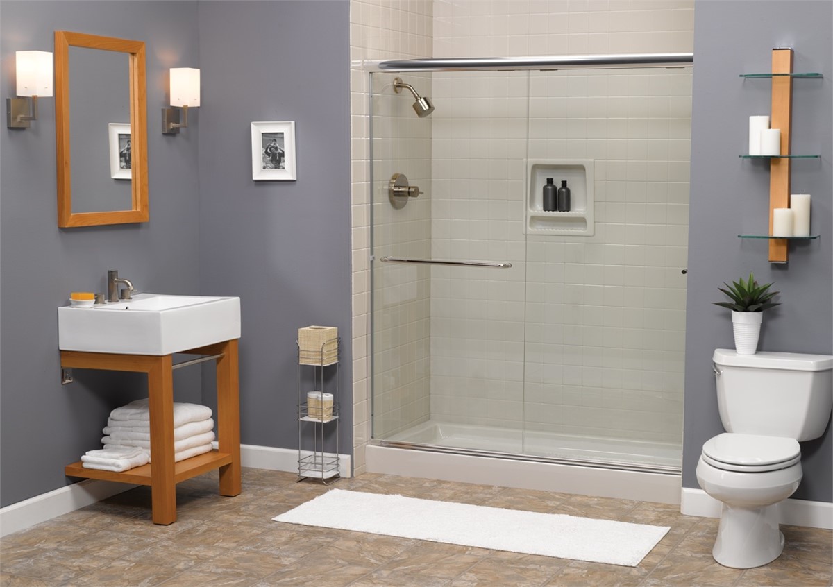 Modern bathroom with a walk-in shower, glass door, gray walls, single sink with wooden accents, toilet, wall shelves with decor, and a white bath mat on the tile floor.