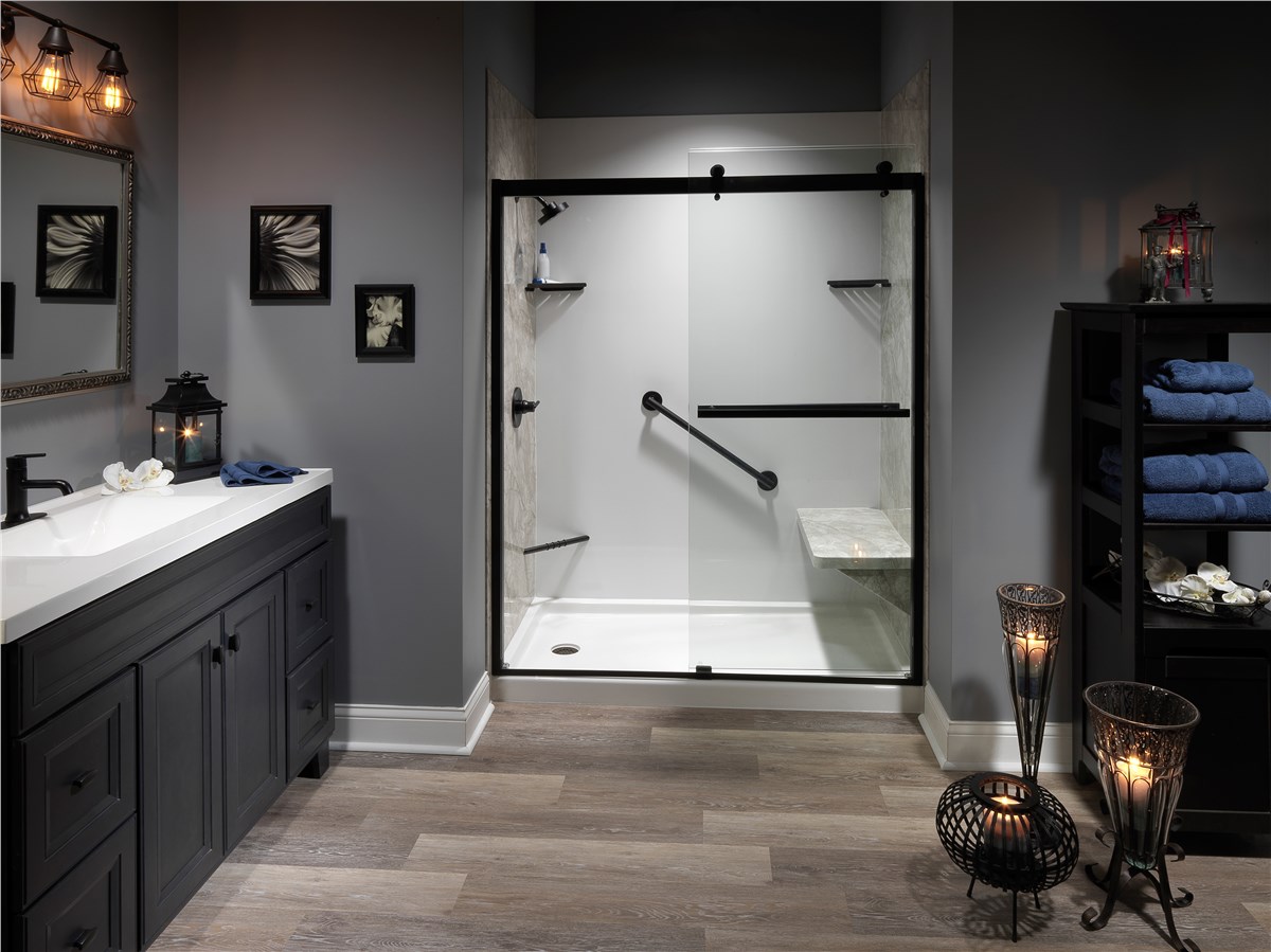 A modern bathroom with dark cabinets, a white countertop, a large walk-in shower with glass doors and black hardware, towel shelves, wall art, lanterns, and wood-style flooring.