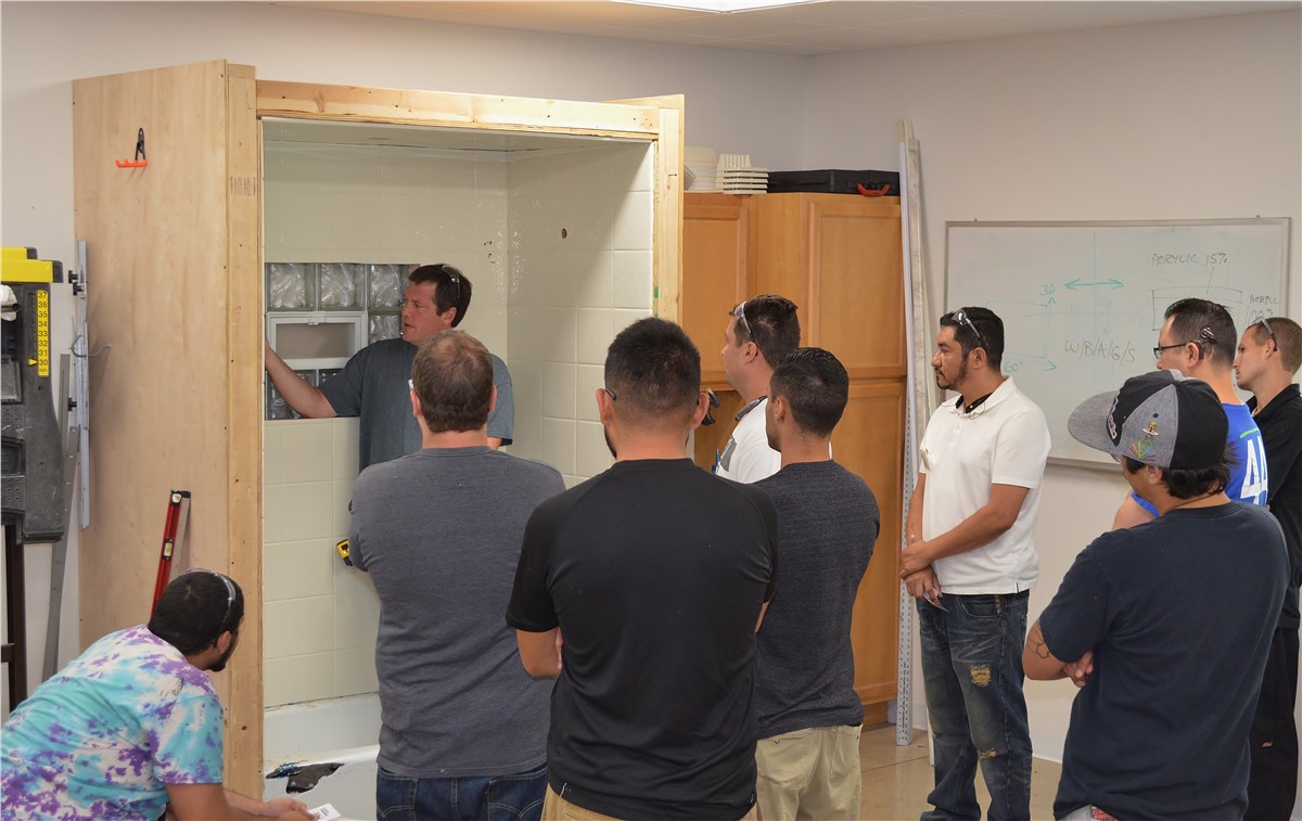 A group of people are gathered around and observing a constructed demonstration of a shower stall within a classroom setting. The shower stall features a tiled interior and glass block window, with a partial wooden frame surrounding it. One man inside the shower stall is gesturing outwards, presumably explaining something to the observers. Several other men stand facing the shower stall, attention focused on the man near the stall. The individuals' expressions are neutral and focused. In the background, a whiteboard covered in diagrams and notes can be partially seen, adding to the classroom setting.
