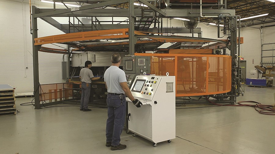 A worker operates a large industrial machine behind a safety barrier, while another worker stands to the left of the machine, and the control panel has a screen and multiple buttons.