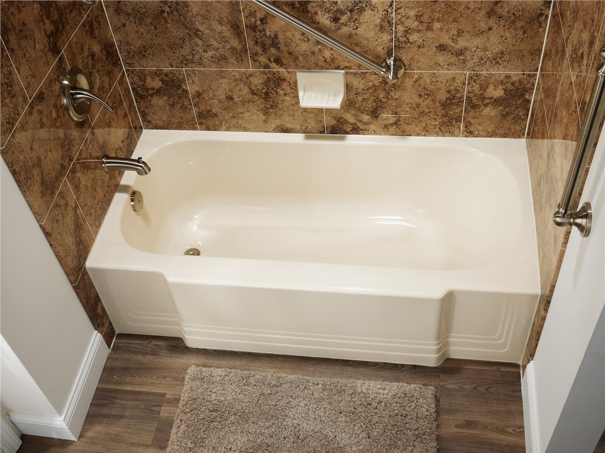 Beige bathtub with chrome handrails affixed to brown tiled wall, above wood-look flooring and a taupe bathmat.