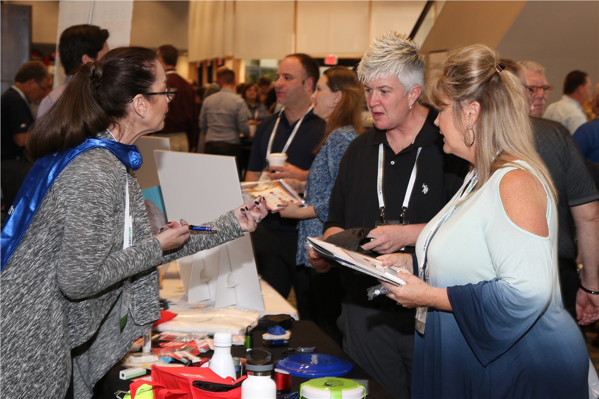 Attendees interact at a conference exhibit table. A woman in a blue cape and lanyard holds out a pen to another woman who holds a brochure. People are looking at brochures and items displayed on the table.