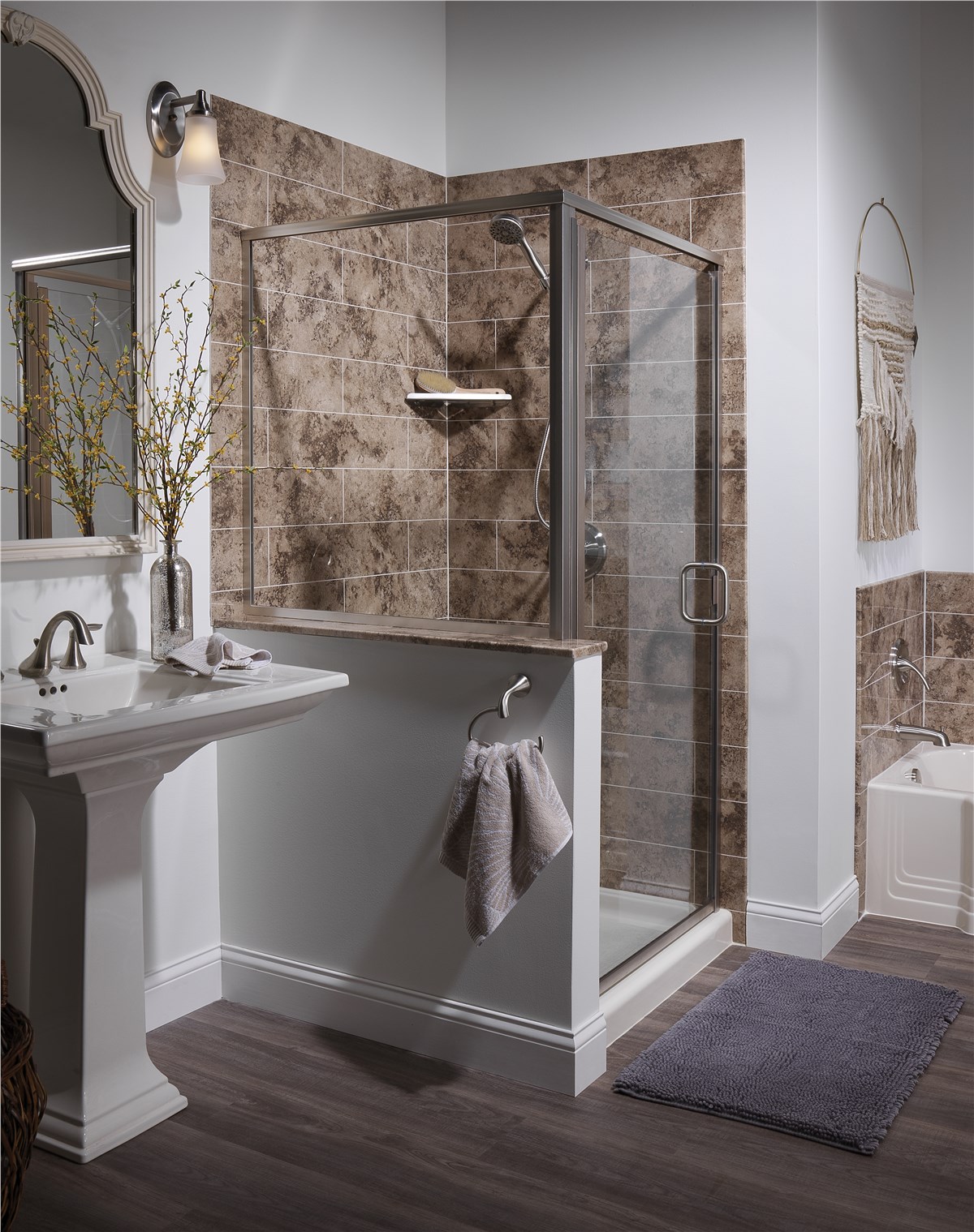 A bathroom with a glass-enclosed shower featuring brown tile walls, a pedestal sink with a mirror, a towel hanging on a hook, a small tub, and a gray bathmat on dark wood flooring.