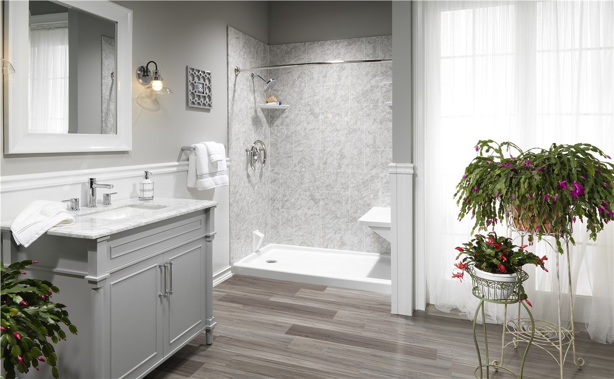 A modern bathroom with gray walls, a large window with sheer curtains, a walk-in shower with gray marble walls, a gray vanity with a marble top, wooden flooring, and two potted flowering plants.