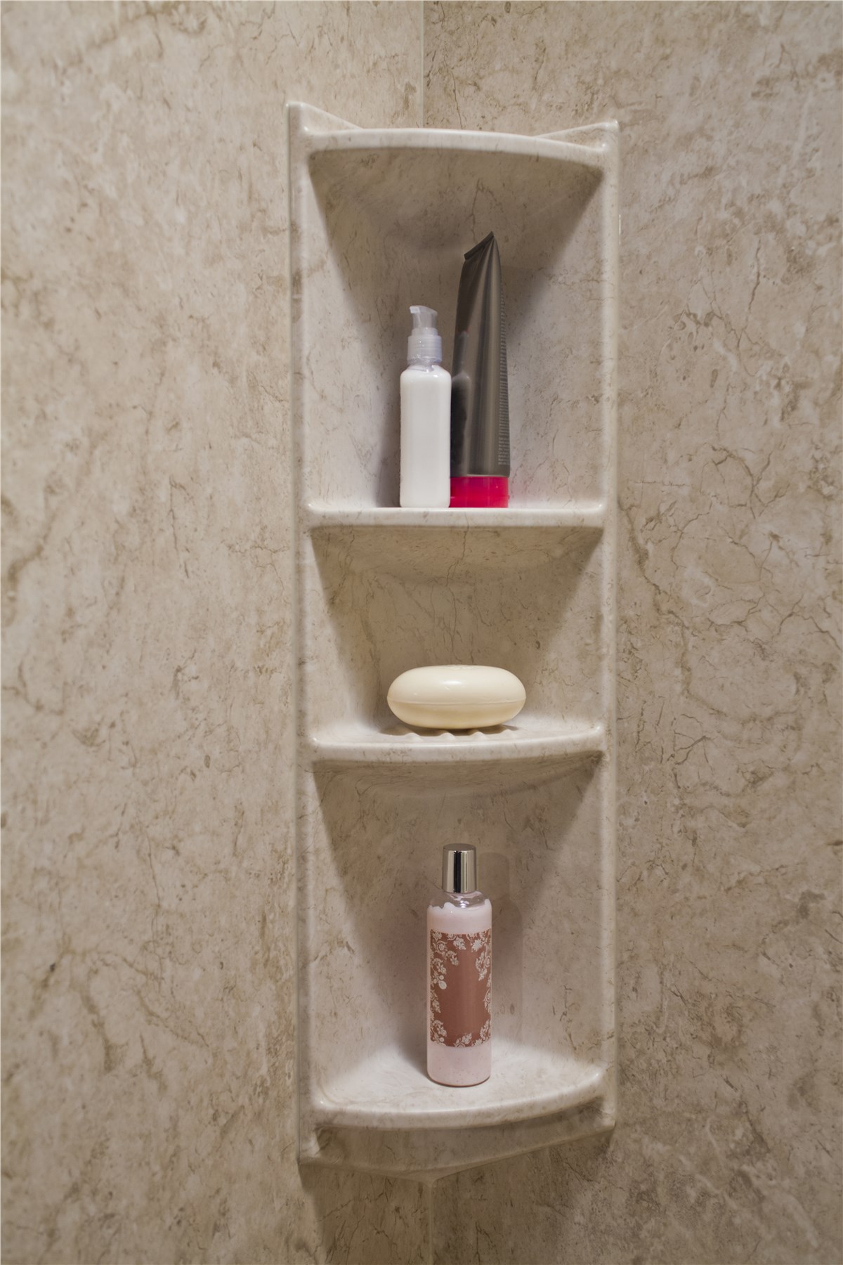 A marble shower corner shelf holds a white pump bottle, a gray bottle with a pink base, an oval bar of soap, and a pink patterned bottle against a beige marble wall.