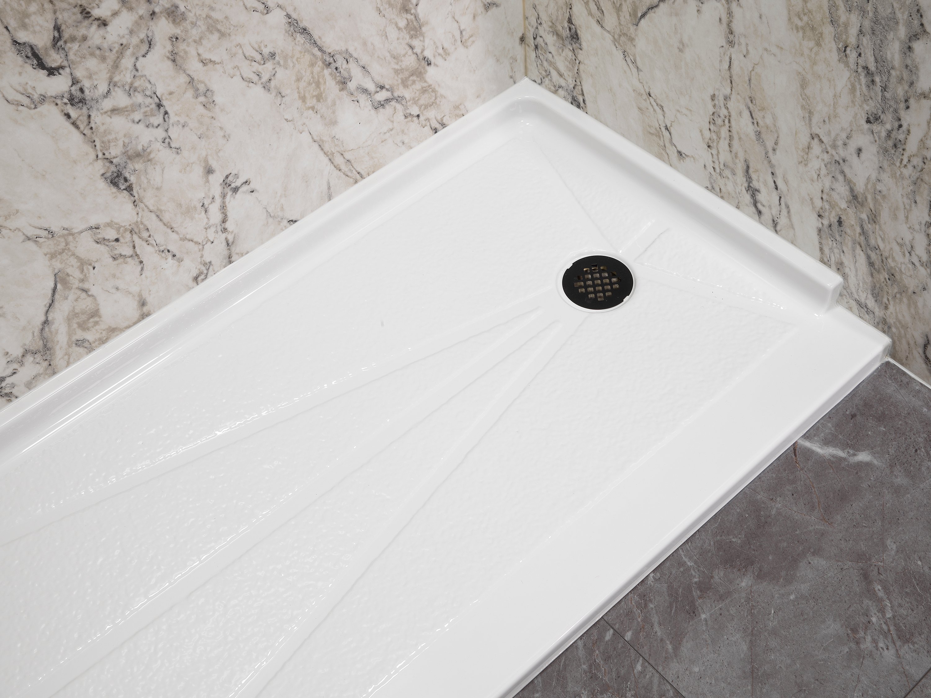 Close-up of a white shower base with a black drain and textured surface, set against marble-patterned wall paneling and a dark gray tile floor.