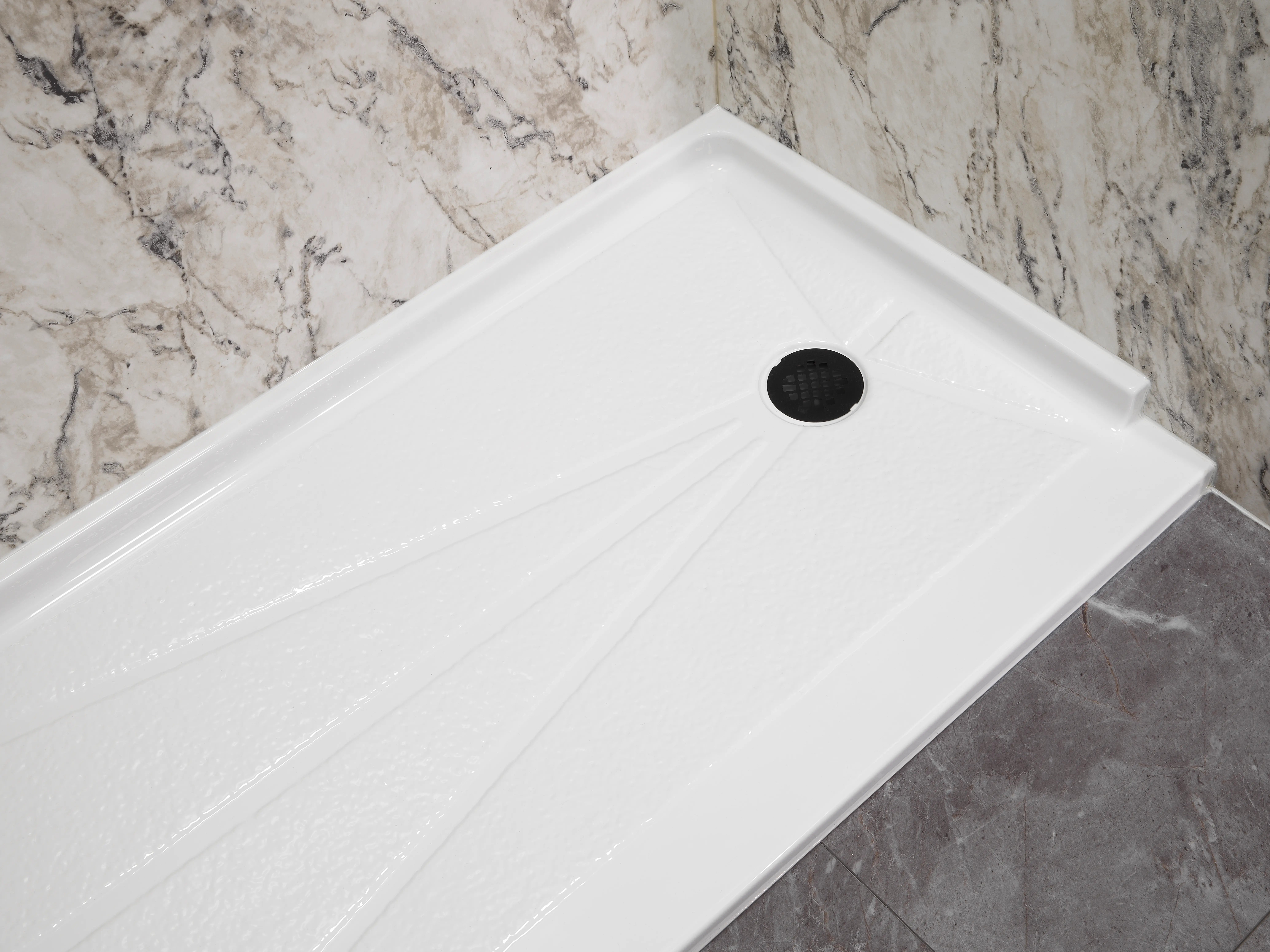 White shower base with a black drain against marble-patterned tiling.