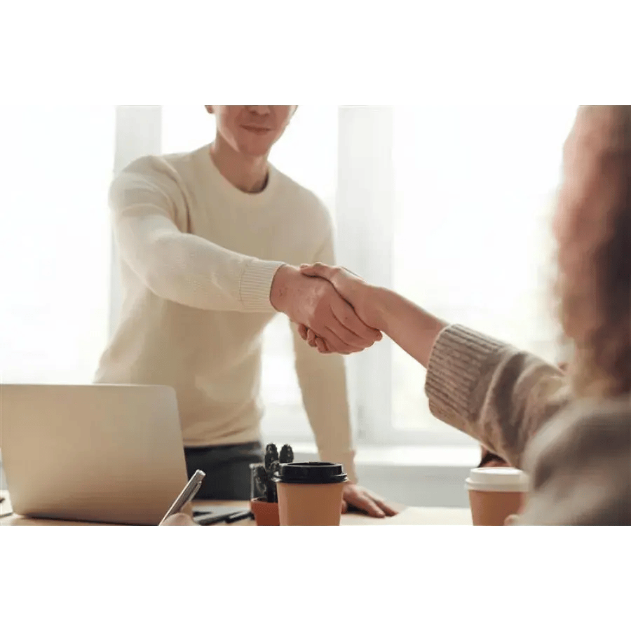 Two people shaking hands over a desk with a laptop, coffee cups, plants, and pens.