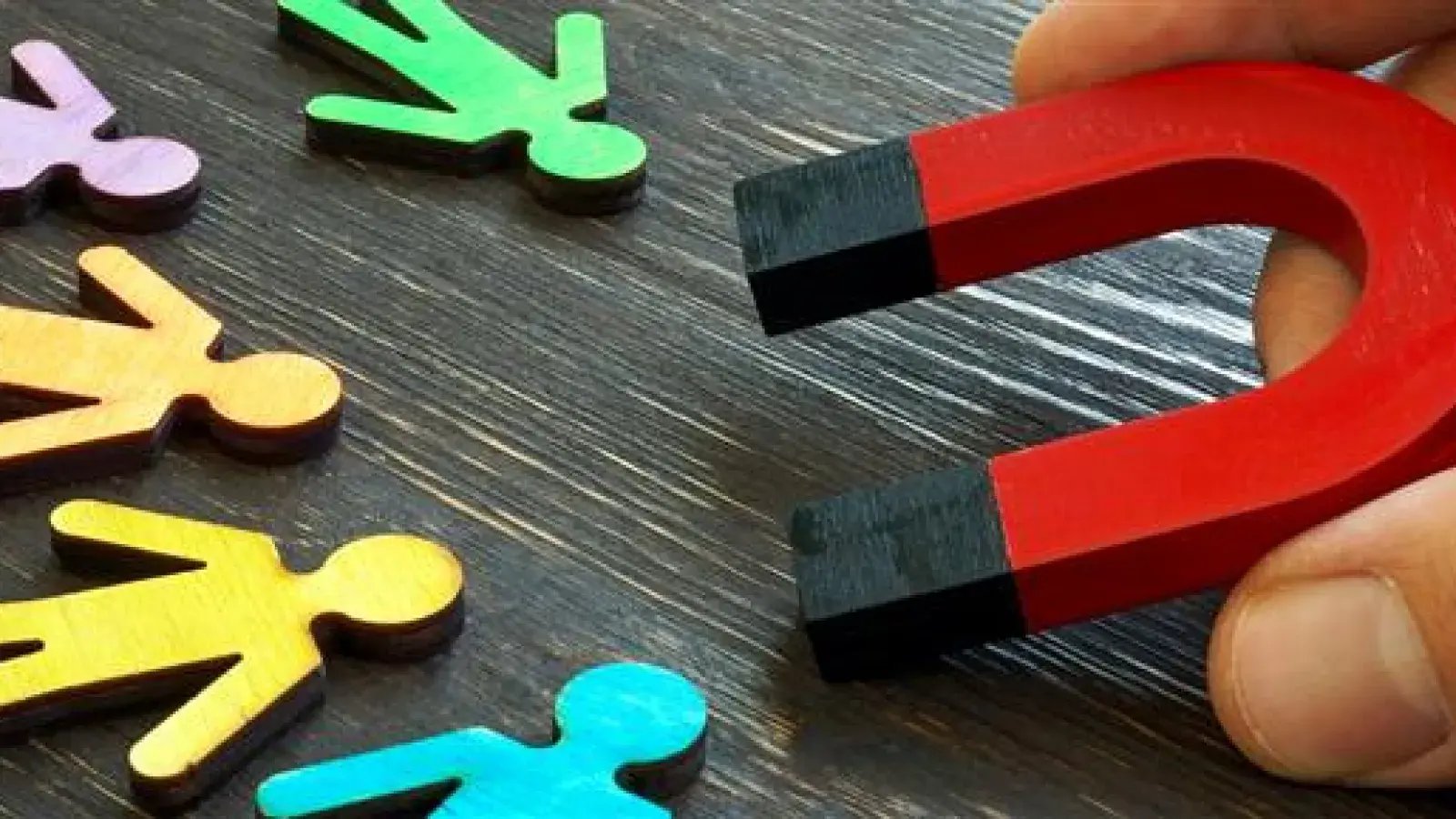 Hand holding a horseshoe magnet near colorful wooden figures on a dark wooden surface.