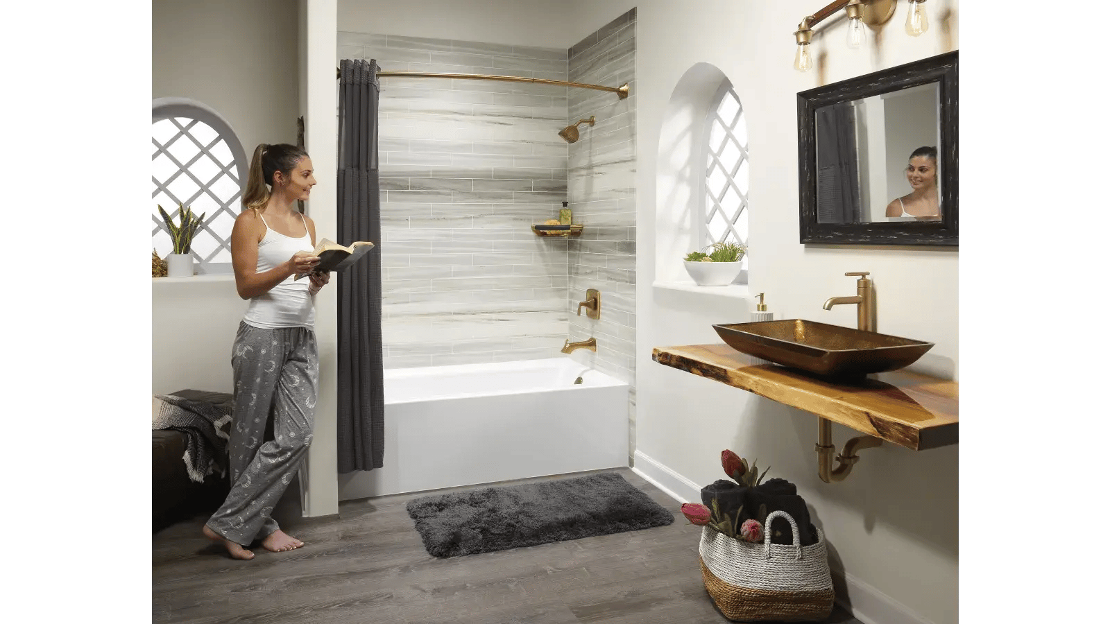 Bathroom featuring a white bathtub with a gray shower curtain surrounded by gray and white horizontal pattern tiles, brass fixtures and shower head, a natural wood countertop with a gold vessel sink, a basket with black towels and pink tulips. A woman with her hair in a ponytail wearing pajamas stands inside the bathroom reading a book as reflected in the mirror.