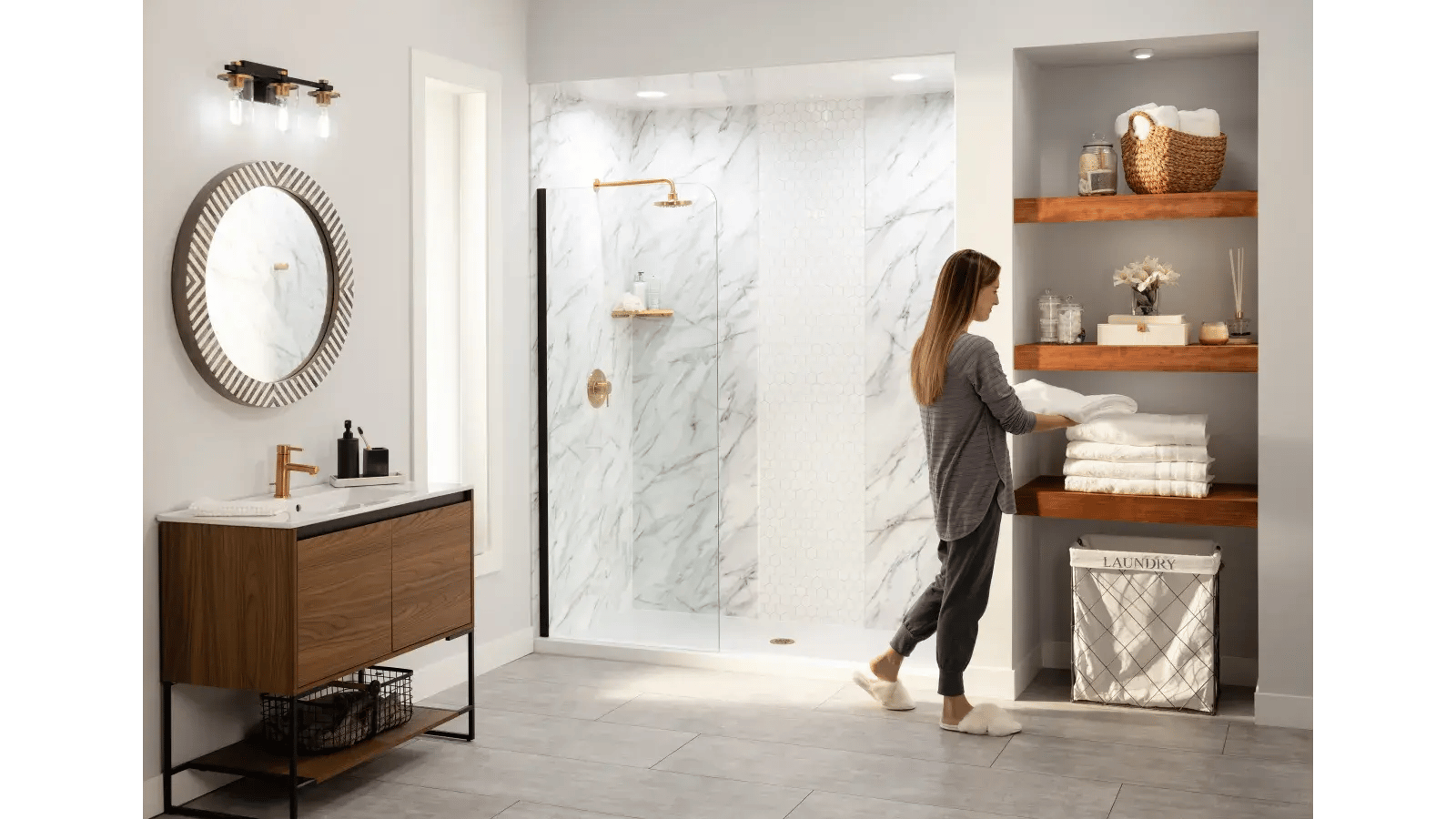 Bathroom featuring wood vanity, round wood-framed mirror, glass enclosed shower with gold fixtures, and woman in pajamas placing white towels on wood shelves built into a niche.