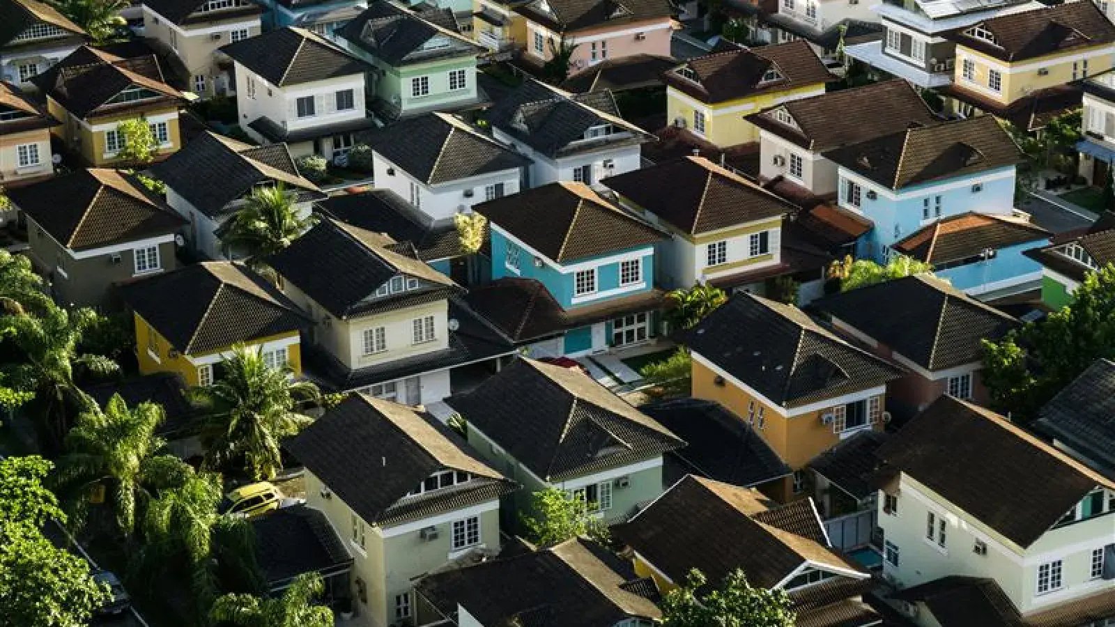 Aerial view of closely packed single-family houses with dark roofs, light painted walls, and green trees interspersed.