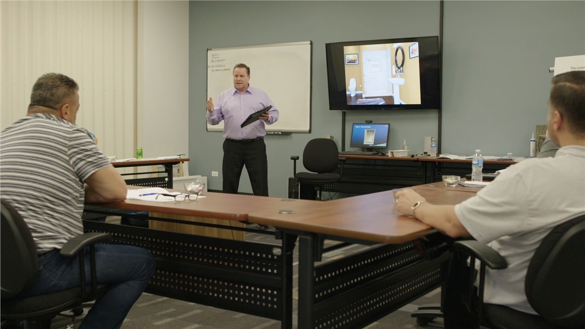 Classroom setting: a male presenter stands before seated attendees, holding a tablet, with a whiteboard to his left and a display showing a bathroom interior on his right.