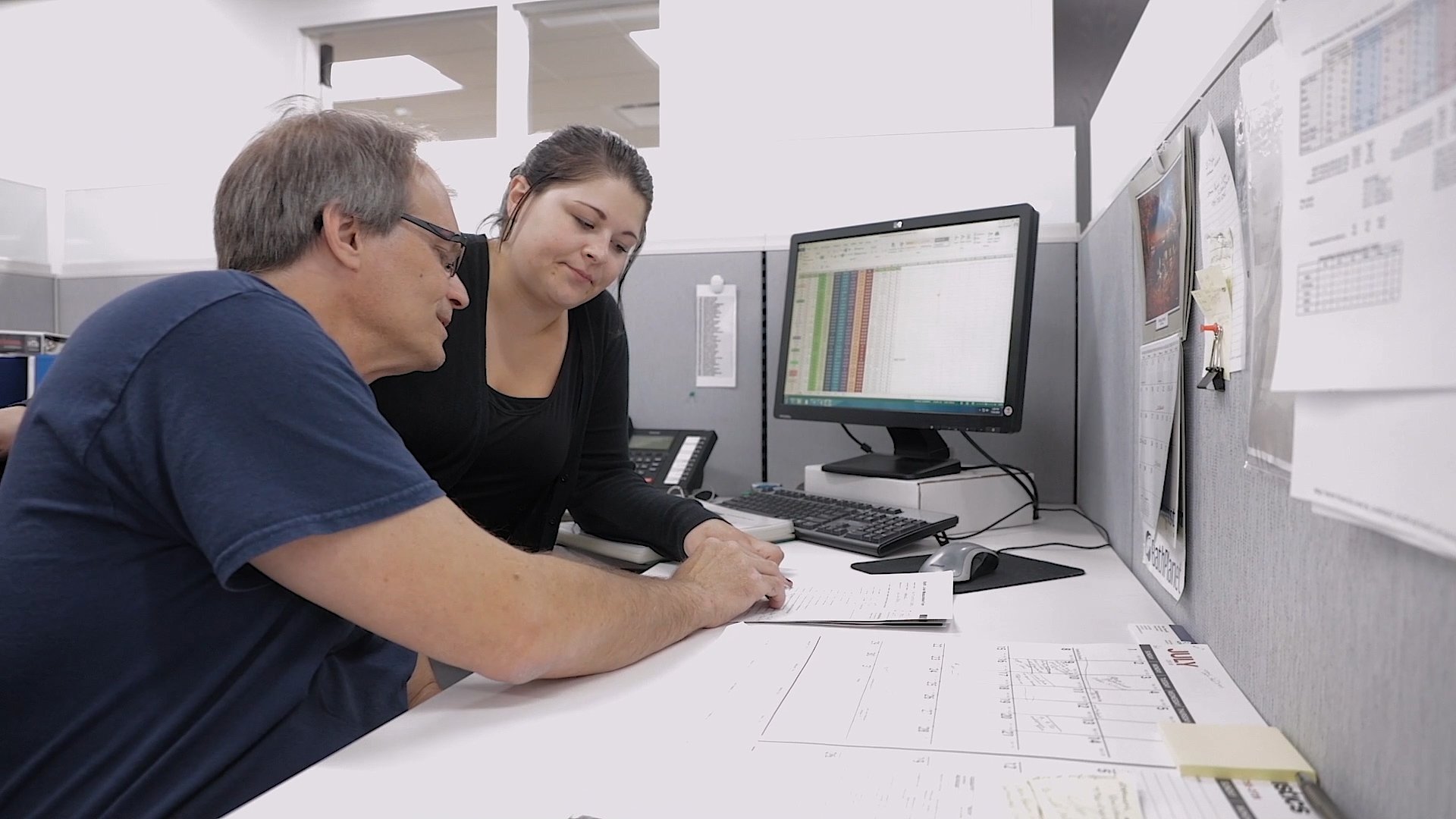 two professionals collaborate at a desk, a man in a blue shirt and glasses is looking with gentle attentiveness at a woman with long dark hair, wearing a black shirt, as she reviews paperwork.