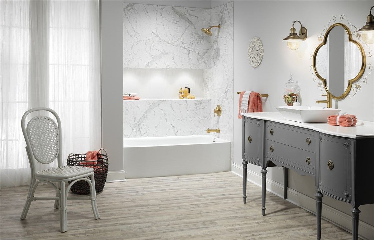 Bathroom with a white tub surrounded by white marble patterned walls, gold fixtures, light gray wooden floor, and a gray vanity with white marble counter.