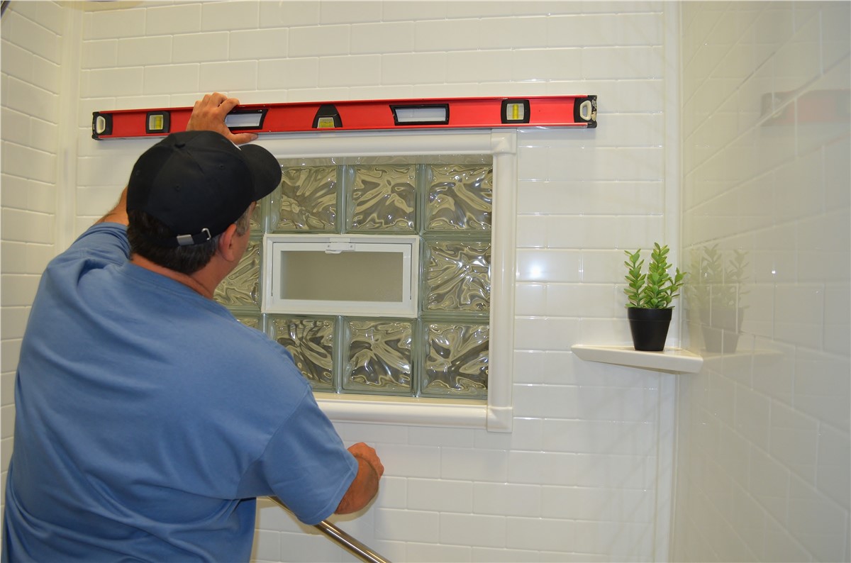 Person using a level to install a glass block window in a white tiled shower, with a small potted plant on a corner shelf.