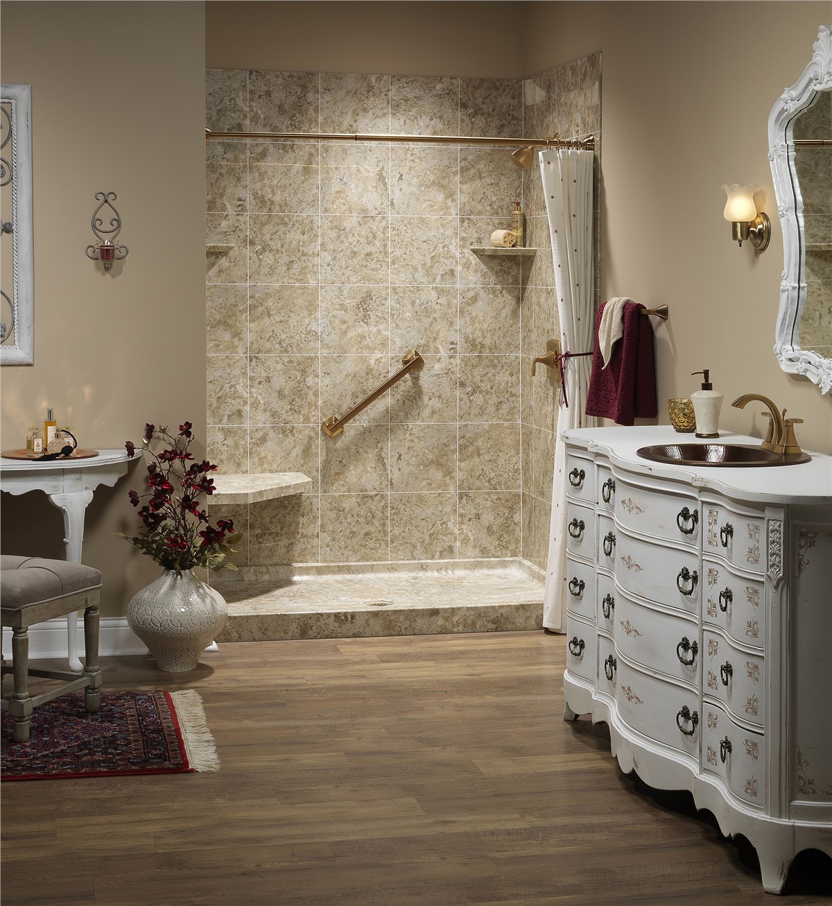A bathroom with a walk-in tiled shower, gold grab bar and seat, a patterned curtain, a white vintage vanity with a mirror, a wall sconce, a white side table, and a vase with flowers on a rug.