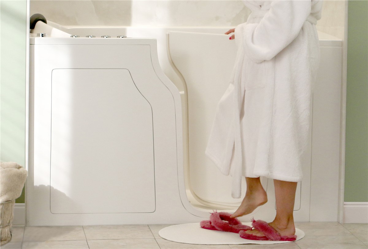 Person in a white robe preparing to enter a walk-in bathtub with a door, stepping from a circular bathmat with fuzzy pink slippers.
