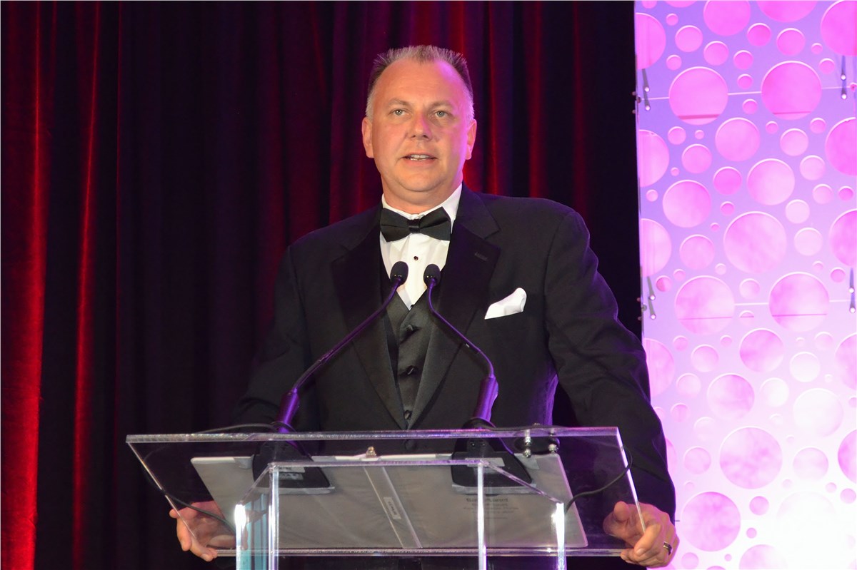 Man in tuxedo standing at a podium, speaking to an unseen audience.