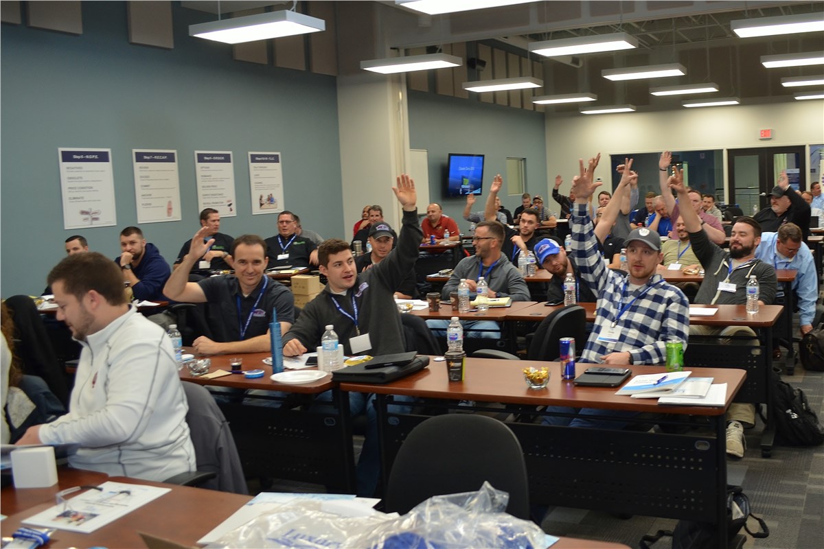 A classroom of approximately 30 adults raises their hands to answer a question during a seminar. Most are wearing casual attire and lanyards. Desks are placed in rows and many have water bottles on them. Display posters are on the light blue walls, while fluorescent lights illuminate the room.