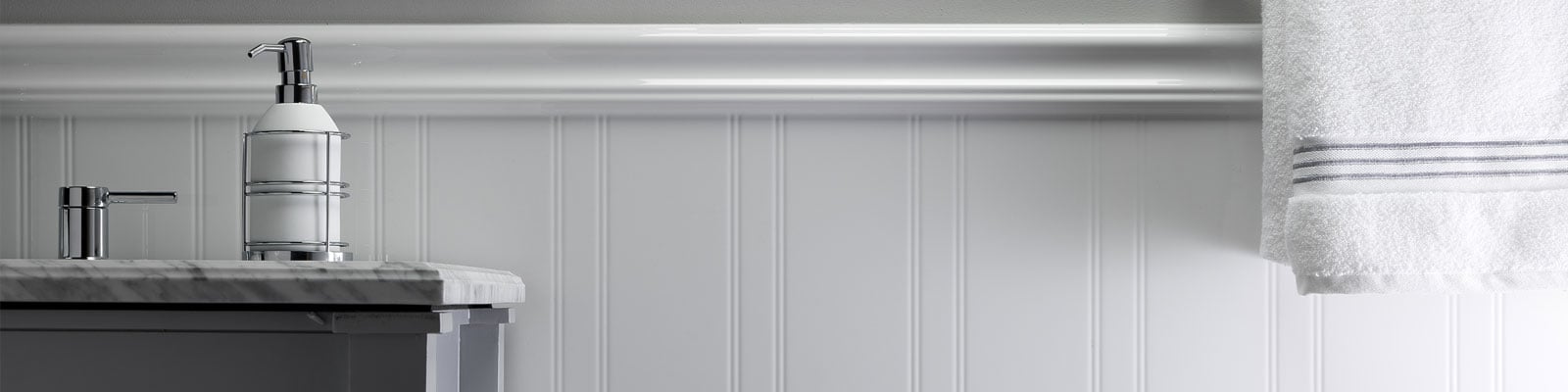 A bathroom countertop with a soap dispenser next to a faucet sits on the left, while a white towel with gray stripes hangs on a wall-mounted rack to the right. The background features white paneled walls.