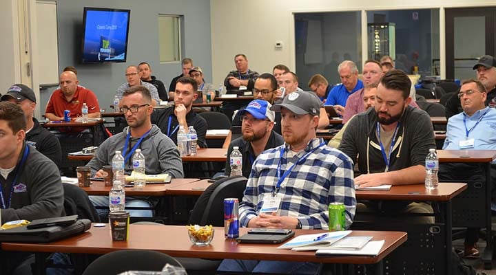 Indoor setting shows rows of adults seated at desks, facing forward towards a monitor. Their facial expressions denote engagement and concentration as they appear to be taking part in a lecture or seminar. Several water bottles, cans and cups are set on the desks.